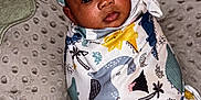 Tyriq is registered to the contest to win money with this photo: baby, infant, swaddle, dinosaur_pattern, cushion, pillows, checkered, face, headwear, lying_down, indoor, child, cute, soft_texture, blanket, wearing, young, adorable, resting, portrait