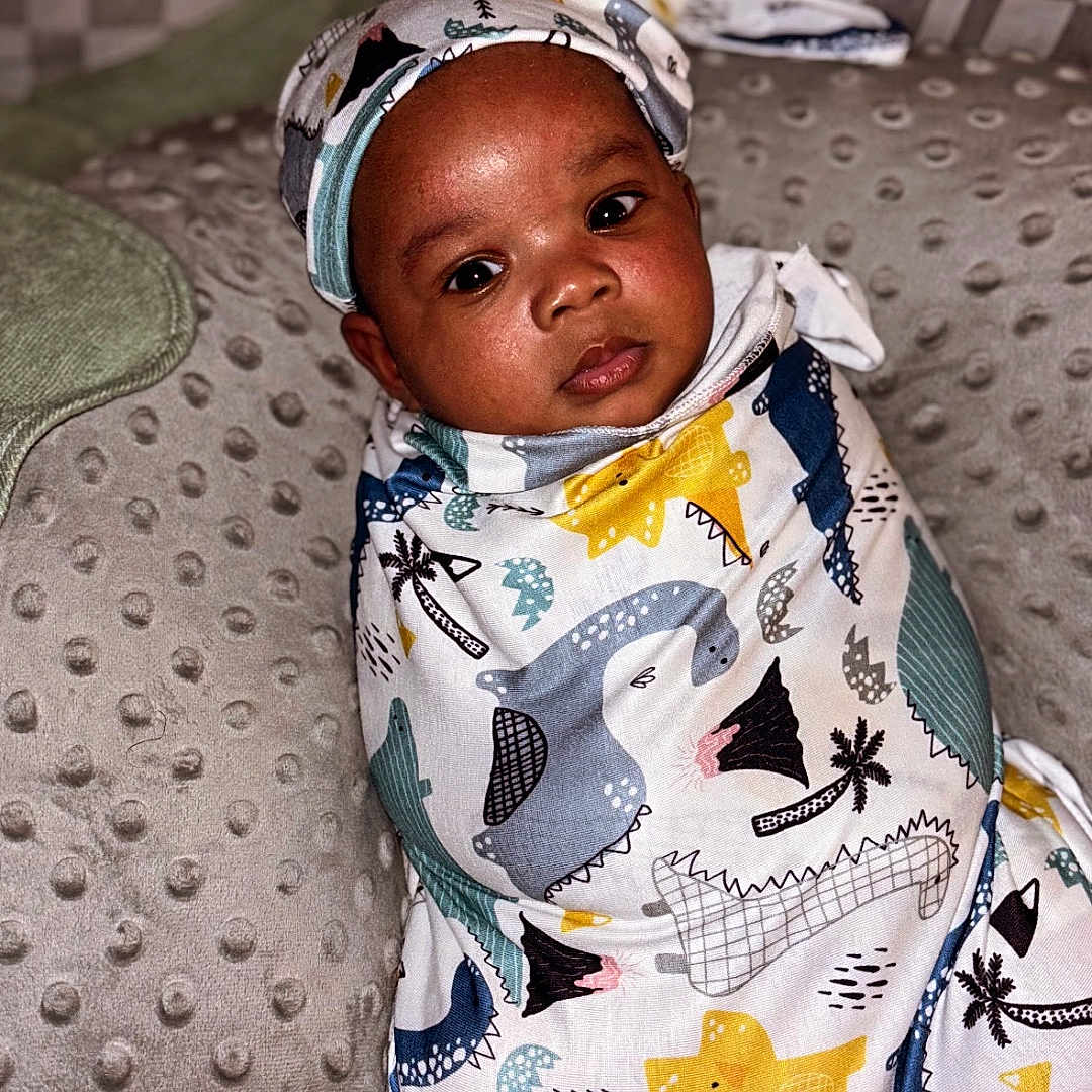 Tyriq is registered to the contest to win money with this photo: adorable, baby, blanket, checkered, child, cushion, cute, dinosaur_pattern, face, headwear, indoor, infant, lying_down, pillows, portrait, resting, soft_texture, swaddle, wearing, young