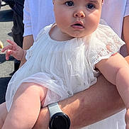 Victoire a rejoint le concours — aidez-le/la à gagner de superbes lots ! baby, child, infant, headband, white_dress, arm, watch, person, outdoor, sunlight, curious, holding, adult, face, skin, clothing, portrait, people, daylight, expression
