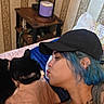 animal, bed, black_cat, blue_hair, cap, cat, closeup, collar, earring, face, indoor, kiss, lamp, person, pet, quilt, resting, tattoo, tattooed_person, wallpaper