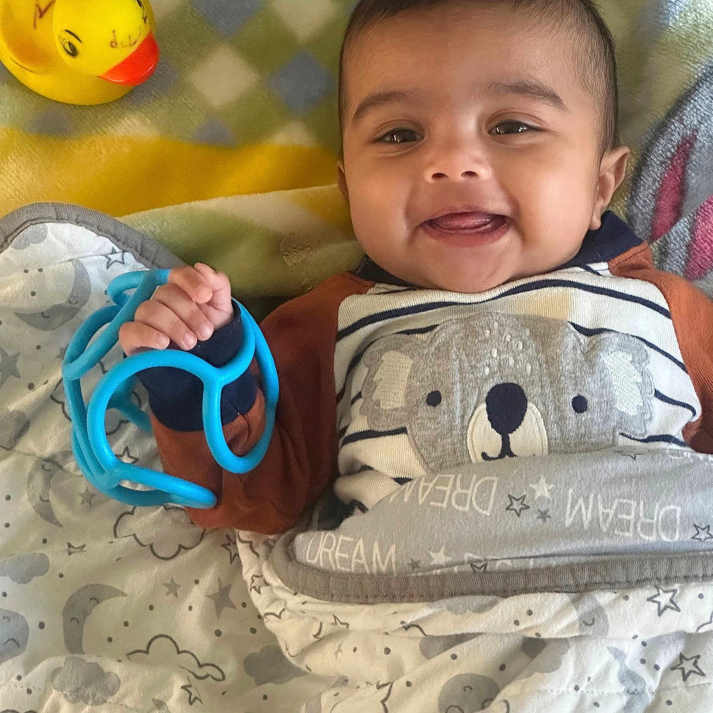 Jovina joined the competition — help win amazing prizes! baby, infant, smiling, happy, portrait, lying_down, toy, rubber_duck, blue_toy, blanket, cozy, bed, bear_outfit, striped_clothing, patterned_fabric, nursery, indoors, hand, face, close_up