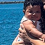 Khamari is registered to the contest to win money with this photo: baby, child, person, curly_hair, water, outdoor, holding, adult, arm, hand, blue_sky, sea, coast, rail, clothing, white_outfit, portrait, cute, sunlight, daytime
