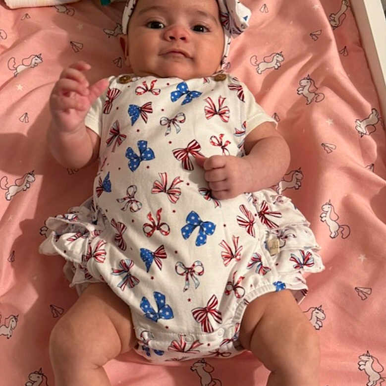 Celestial is registered to the contest to win money with this photo: baby, baby_accessories, blanket, blue_bows, child, cute, face, hand_raised, headband, indoor, infant, lying_down, onesie, person, pink, red_bows, sleeping_area, soft_toys, unicorn_pattern, white_clothing