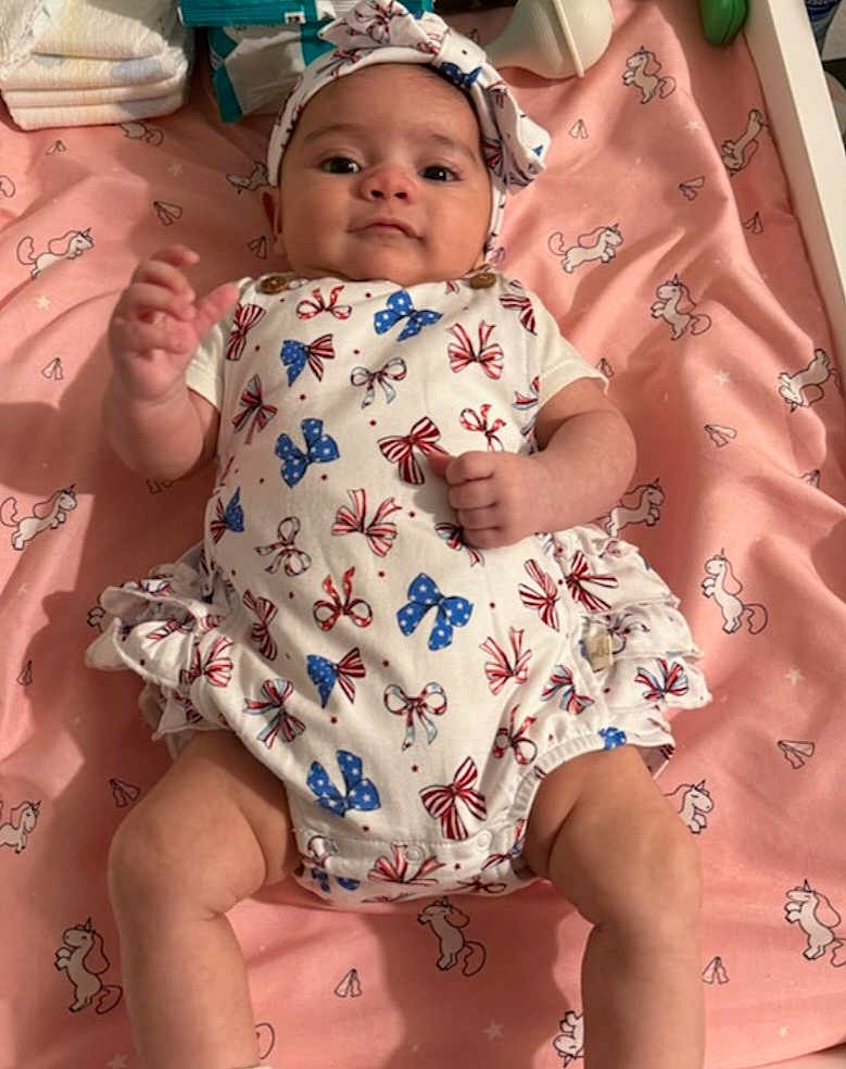 Celestial is registered to the contest to win money with this photo: baby, infant, onesie, headband, blanket, unicorn_pattern, pink, white_clothing, red_bows, blue_bows, lying_down, hand_raised, indoor, cute, child, face, person, soft_toys, sleeping_area, baby_accessories