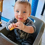 Anthony is registered to the contest to win money with this photo: baby, child, high_chair, smile, happy, snack, camo_clothing, indoor, play_area, net, blue_padded_pole, table, hands, face, person, cute, toddler, joyful, seated, pajamas