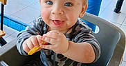 Anthony is registered to the contest to win money with this photo: baby, blue_padded_pole, camo_clothing, child, cute, face, hands, happy, high_chair, indoor, joyful, net, pajamas, person, play_area, seated, smile, snack, table, toddler