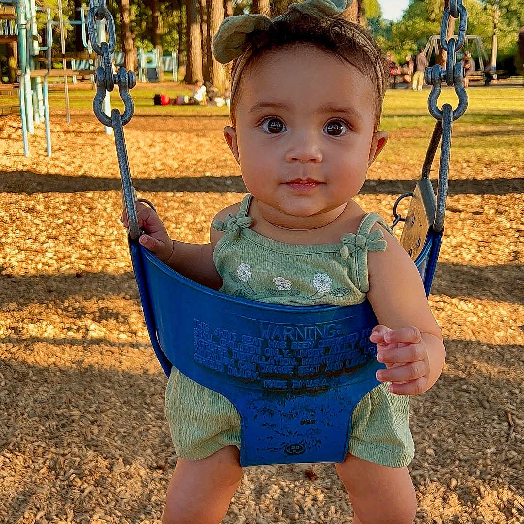 Carolina is registered to the contest to win money with this photo: baby, barefoot, bow, child, cute, daytime, fun, green_outfit, infant, outdoor, park, person, play, playground, smiling, summer, sunlight, swing, trees, woodchips