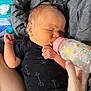 baby, newborn, bottle, feeding, black_onesie, arrow_pattern, blanket, gray_blanket, hand, arm, infant, sleeping, closeup, skin, person, baby_bottle, pink_cap, resting, cozy, indoor