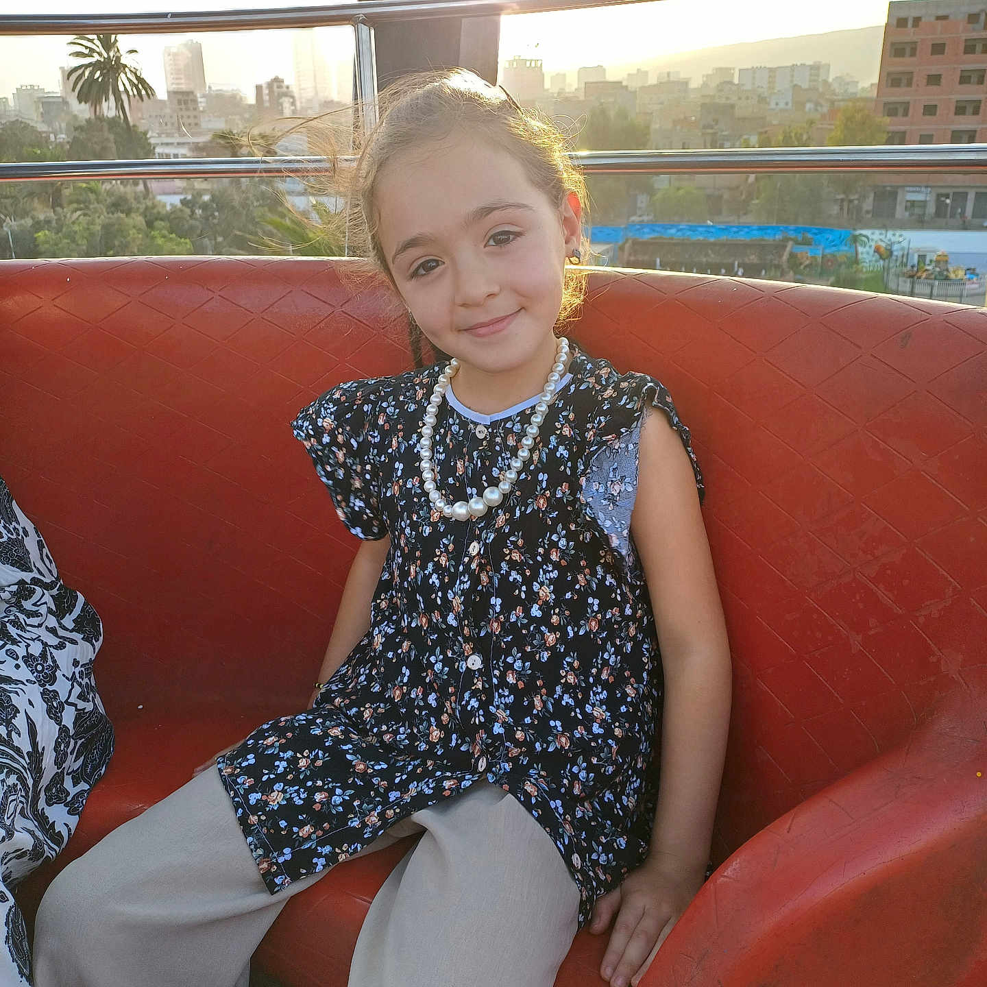 Selma a rejoint le concours — aidez-le/la à gagner de superbes lots ! casual, child, cityscape, daytime, fashion, floral_top, girl, happy, nature, outdoor, pants, pearl_necklace, portrait, railing, red_bench, relaxed, sitting, smile, sunlight, urban