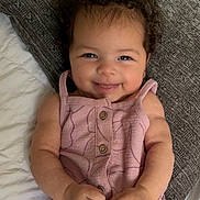 Capri joined the competition — help win amazing prizes! baby, child, smiling, curly_hair, pink_clothing, romper, pillow, bed, texture, cute, happy, infant, skin, hands, face, eyes, indoor, portrait, lying_down, young