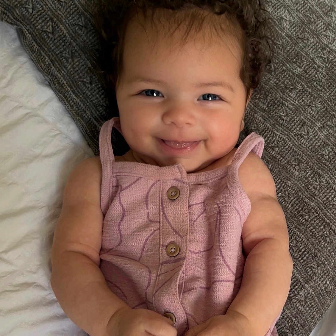 Capri joined the competition — help win amazing prizes! baby, bed, child, curly_hair, cute, eyes, face, hands, happy, indoor, infant, lying_down, pillow, pink_clothing, portrait, romper, skin, smiling, texture, young