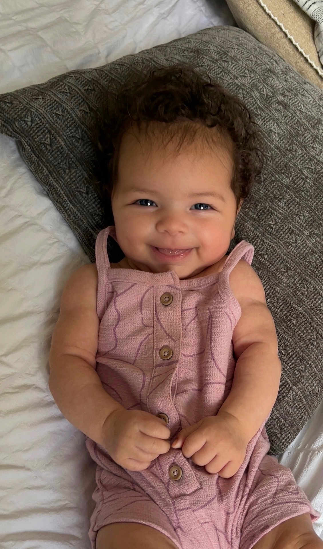 Capri joined the competition — help win amazing prizes! baby, child, smiling, curly_hair, pink_clothing, romper, pillow, bed, texture, cute, happy, infant, skin, hands, face, eyes, indoor, portrait, lying_down, young