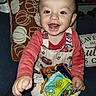 baby, child, infant, smiling, happy, sitting, portrait, indoors, sofa, couch, cushion, toy, book, colorful, overalls, clothing, face, hands, living_room, photograph