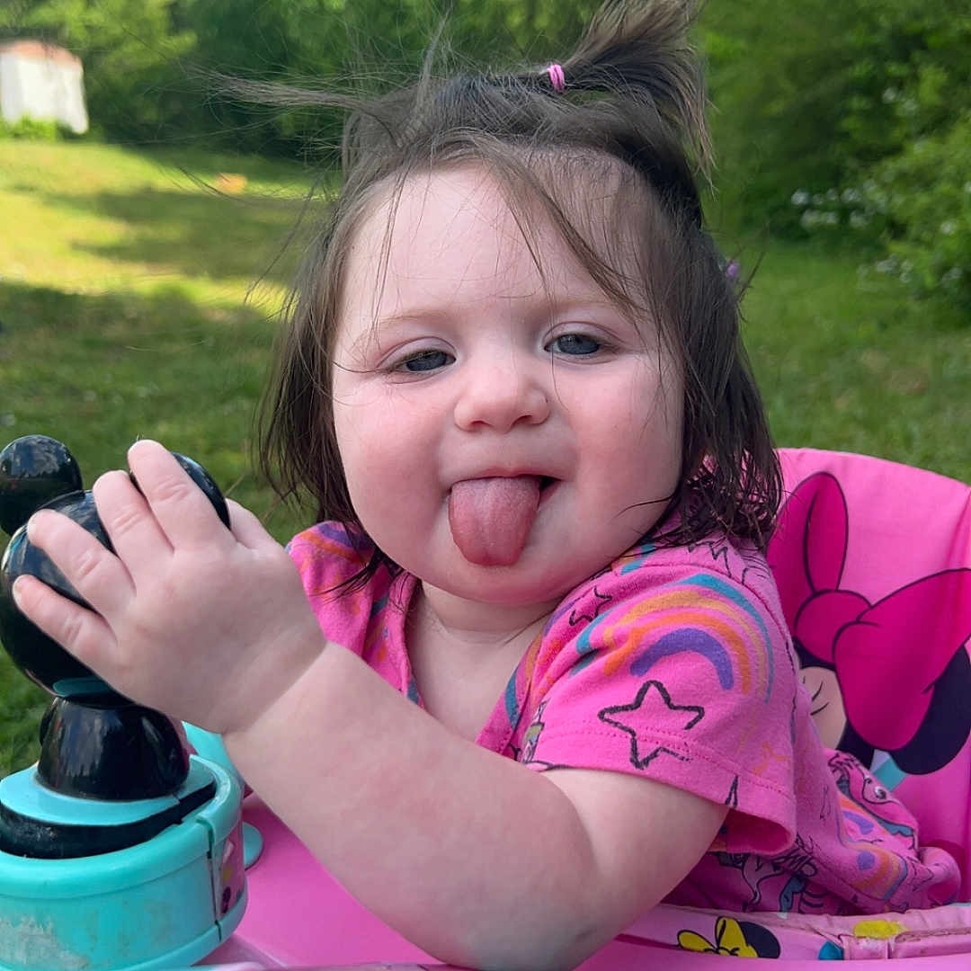 Scarlett is registered to the contest to win money with this photo: toddler, child, playful, tongue_out, pink_clothing, toy, walker, outdoor, greenery, grass, sunlight, hair_tie, baby_toy, happy, cute, portrait, summer, nature, fun, smiling