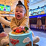 baby, cake, birthday_cake, child, smiling, hands, sweater, indoor, restaurant, bar, bar_stools, blue_lights, wooden_pillar, tv_screen, celebration, toy_car, frosting, portrait, joyful, person