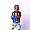 black_shoes, black_tshirt, blue_ball, boy, casual_clothing, child, cute, denim_shorts, footwear, happy, indoor, orange_socks, playful, portrait, shorts, smiling, standing, studio, toddler, white_background