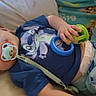 baby, pacifier, toy, blue_shirt, cartoon_character, pants, sailboat_pattern, blanket, lying_down, infant, child, cute, indoors, comfort, soft_surface, playful, colorful, hand, face, sleepwear