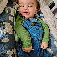 Mateo is registered to the contest to win money with this photo: baby, infant, smiling, denim_overalls, green_shirt, pacifier, blanket, cozy, indoor, cute, child, face, happy, lying_down, clothing, closeup, person, soft_texture, home, portrait