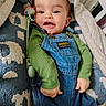 baby, infant, smiling, denim_overalls, green_shirt, pacifier, blanket, cozy, indoor, cute, child, face, happy, lying_down, clothing, closeup, person, soft_texture, home, portrait