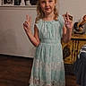 child, girl, peace_sign, dress, indoor, wood_floor, smile, home_decor, photo_frame, rooster_statue, wall_art, furniture, sleeveless_dress, sandals, happy, person, standing, young, casual, light