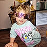 toddler, child, sunglasses, pink, cartoon_tshirt, indoor, cat, black_and_white_cat, wooden_floor, kitchen, cabinet, flower_vase, yellow_flowers, hand, person, smiling, curly_hair, home_interior, furniture, decorative_rooster