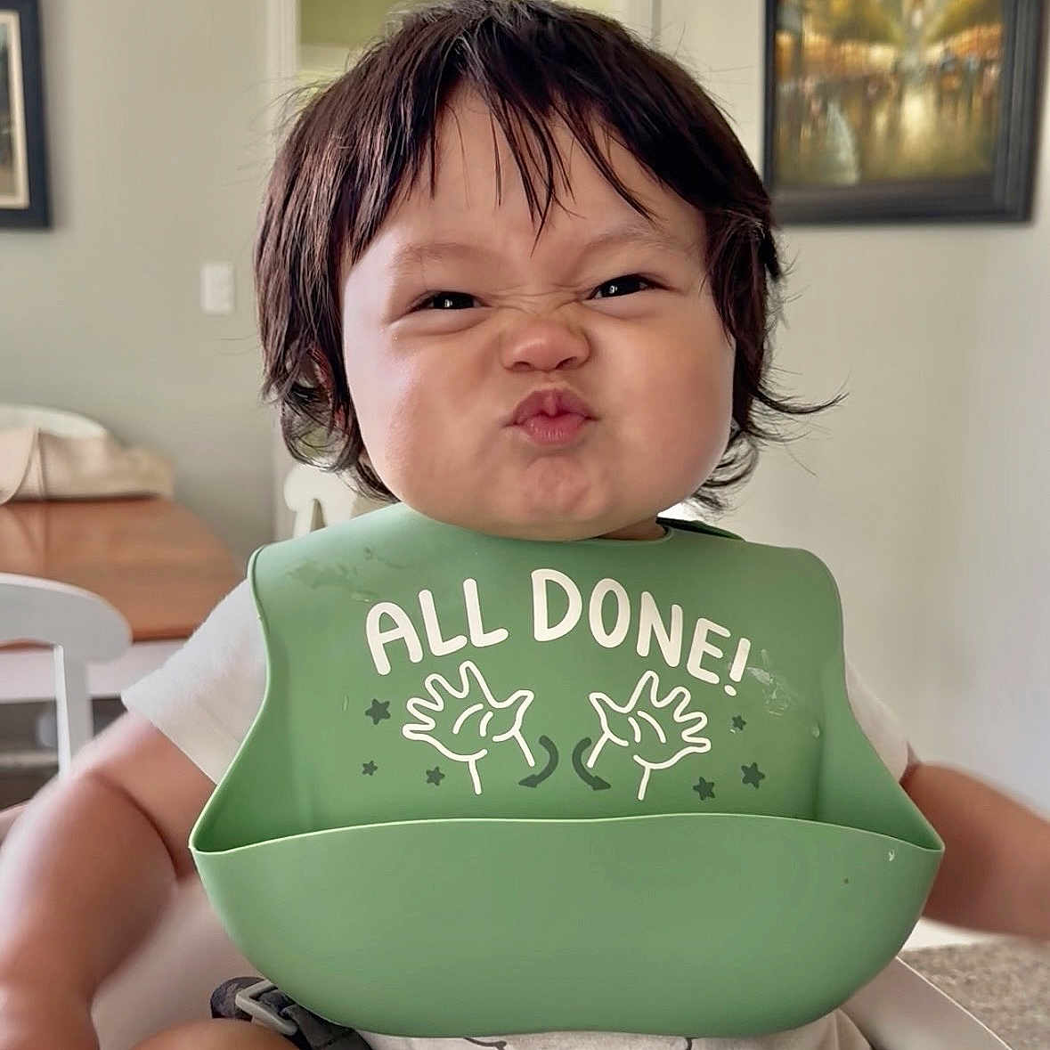 Nils is registered to the contest to win money with this photo: baby, child, high_chair, green_bib, funny_face, indoor, home, dining_table, painting, short_hair, white_shirt, cute, infant, feeding_bib, expression, person, smile, seated, room, furniture