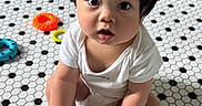 Nils is registered to the contest to win money with this photo: baby, child, floor, tiles, white_clothing, sitting, indoor, toy, colorful, foot, hand, curly_hair, expression, looking_up, skin, playtime, cute, person, young_child, floor_pattern