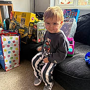 Tommy is registered to the contest to win money with this photo: toddler, child, indoor, sofa, toy, gift_box, window, carpet, clothing, sneakers, striped_pants, sweatshirt, face, smile, furniture, holiday_decor, santa_sticker, playroom, candid, person
