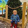 toddler, child, christmas_tree, red_boots, blue_jacket, smiling, indoor, public_space, wooden_fence, marble_floor, holiday_decor, festive, casual_clothing, small_child, curly_hair, seated, happy, cute, lighting, portrait