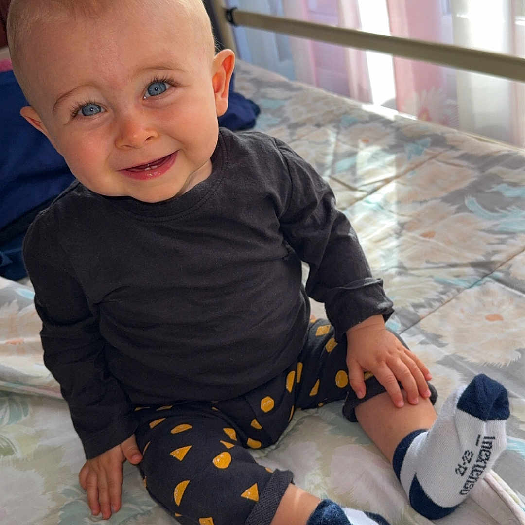 Eden participe au concours pour gagner de l'argent avec cette photo : baby, bed, black_shirt, blanket, blue_eyes, child, clothing, curtains, face, footwear, hand, happy, indoors, natural_light, patterned_pants, portrait, sitting, smiling, socks, toddler