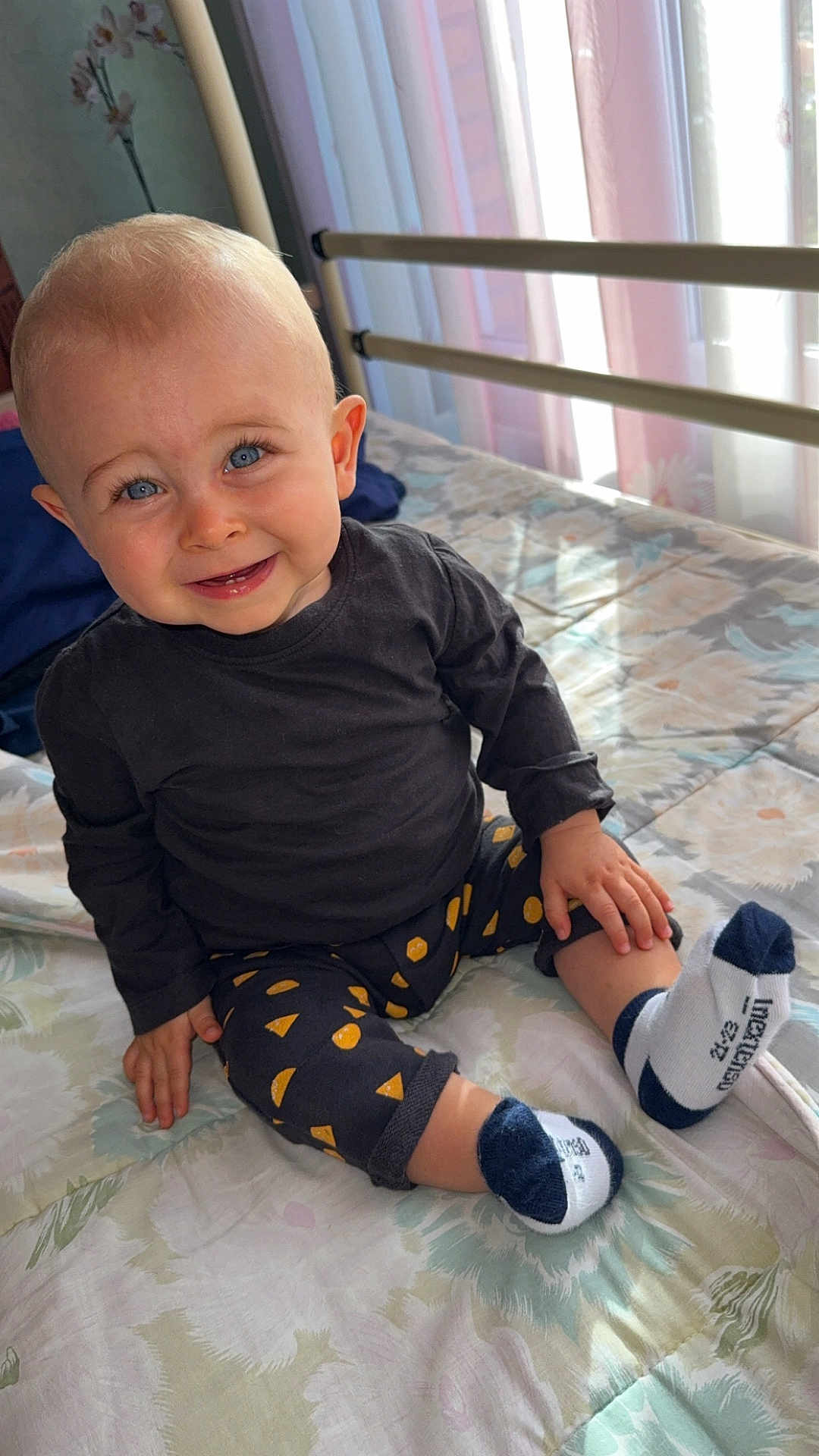 Eden participe au concours pour gagner de l'argent avec cette photo : toddler, child, baby, smiling, blue_eyes, sitting, indoors, bed, blanket, clothing, black_shirt, patterned_pants, socks, happy, curtains, natural_light, face, hand, footwear, portrait