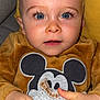 baby, child, blue_eyes, mickey_mouse, sweater, blanket, couch, indoor, closeup, face, hands, cute, soft_texture, cozy, infant, portrait, young_child, expression, holding, furniture