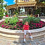 child, toddler, flowerbed, flowers, statue, bench, outdoor, daylight, building, sky, greenery, playful, smile, pavement, shoes, casual_clothing, sculpture, amusement_park, park, sunlight