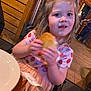 child, girl, bread, food, eating, strawberry_pattern, dress, restaurant, wooden_wall, high_chair, indoor, person, table, plate, cute, smiling, blonde_hair, casual_clothing, dining, happy