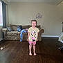 Callie is registered to the contest to win money with this photo: child, smile, indoor, living_room, couch, clothing, floor, window, curtains, tshirt, shorts, barefoot, wall_decor, wood_floor, person, happy, furniture, toy, home, casual