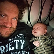 Marshall is registered to the contest to win money with this photo: man, baby, sleeping, smiling, selfie, bed, headboard, onesie, dinosaur_pattern, striped_clothing, indoor, portrait, people, casual, resting, closeup, facial_hair, hair, light, cozy