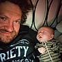 man, baby, sleeping, smiling, selfie, bed, headboard, onesie, dinosaur_pattern, striped_clothing, indoor, portrait, people, casual, resting, closeup, facial_hair, hair, light, cozy