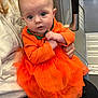 Effie is registered to the contest to win money with this photo: baby, child, orange_dress, sparkle, person, seated, indoor, curious_expression, clothing, adult_hand, face, eyes, head, fabric, sitting, human, cute, infant, portrait, casual