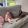baby, dog, couch, blanket, pillow, window, indoor, smiling, pet, child, footwear, tile_floor, greenery, fence, curious, cozy, relaxing, home, light, casual