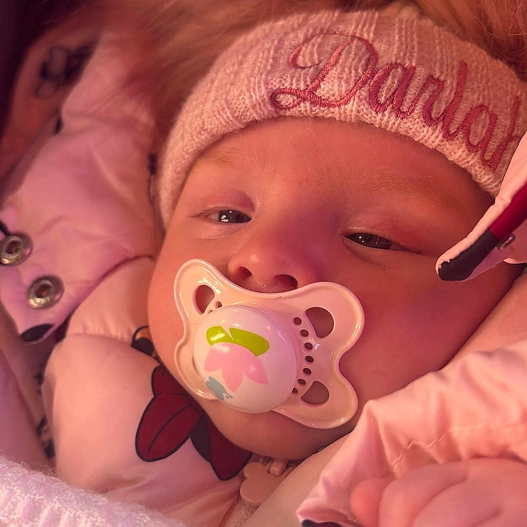 Darlah is registered to the contest to win money with this photo: baby, blanket, child, closeup, cozy, cute, face, fur_trim, hand, hat, infant, pacifier, pink_jacket, portrait, sleepy, snug, soft, warm, winter_clothing, young_child