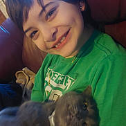 Smokey is registered to the contest to win money with this photo: child, smile, dimple, green_shirt, cat, gray_cat, pet, indoor, couch, leather_sofa, short_hair, necklace, earring, portrait, holding, happy, closeup, living_room, cozy, candid