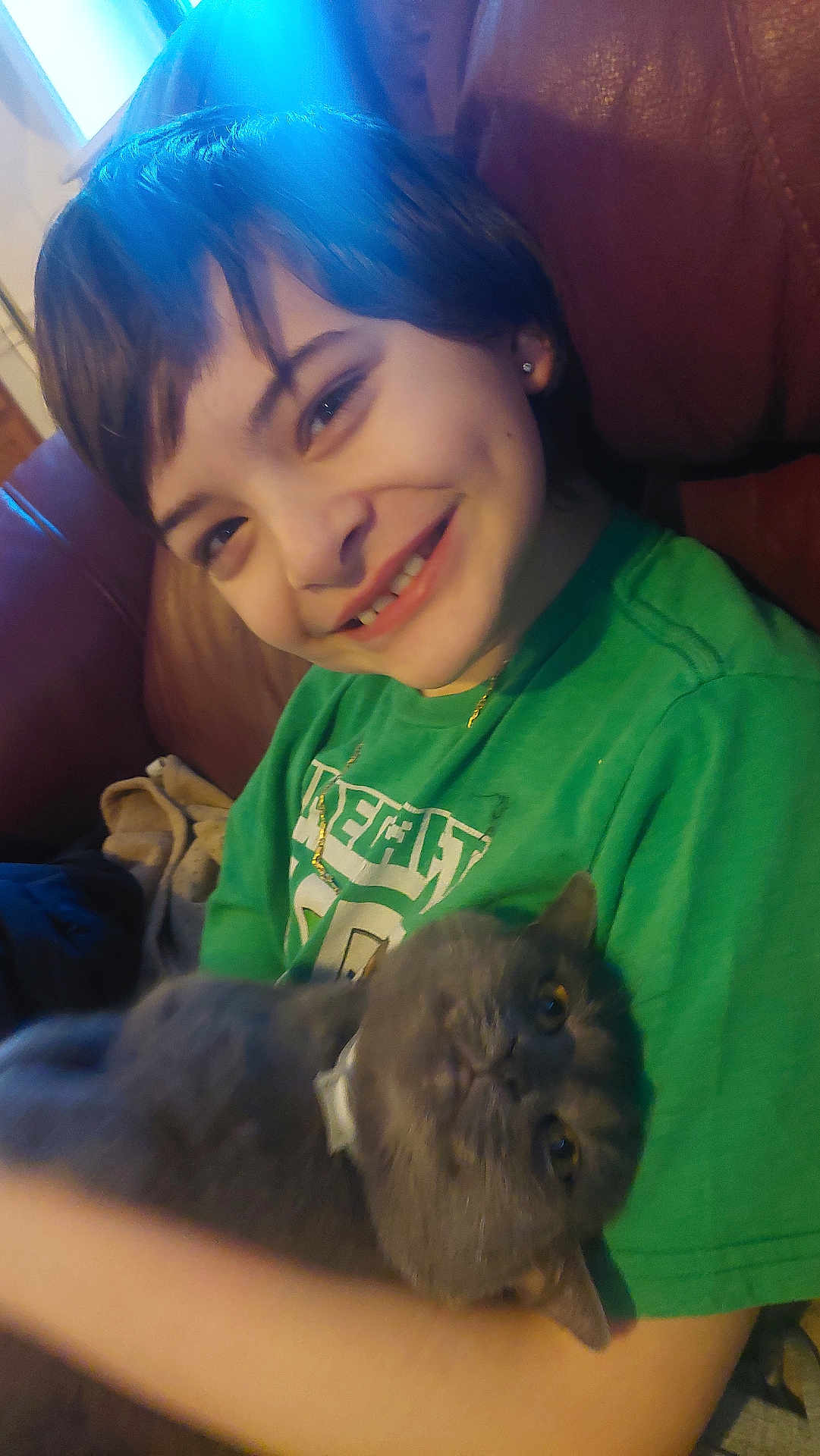Smokey is registered to the contest to win money with this photo: child, smile, dimple, green_shirt, cat, gray_cat, pet, indoor, couch, leather_sofa, short_hair, necklace, earring, portrait, holding, happy, closeup, living_room, cozy, candid