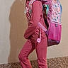 child, girl, pink_outfit, backpack, sneakers, carpet, wall, pose, standing, indoor, smile, hair_ties, casual_clothing, footwear, young, person, playful, accessories, school_item, portrait