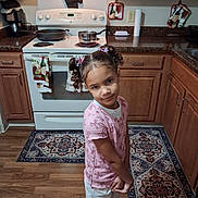 Myra joined the competition — help win amazing prizes! child, girl, portrait, smiling, standing, kitchen, stove, oven, cookware, pots, pans, cabinets, wooden_floor, rug, countertop, kitchen_towels, hair_bows, pink_shirt, white_pants, indoor