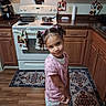 child, girl, portrait, smiling, standing, kitchen, stove, oven, cookware, pots, pans, cabinets, wooden_floor, rug, countertop, kitchen_towels, hair_bows, pink_shirt, white_pants, indoor