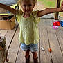 Bella is registered to the contest to win money with this photo: child, yellow_top, denim_shorts, brown_boots, wooden_porch, red_pickup_truck, outdoor, smiling, curly_hair, toy_cars, cardboard_box, green_grass, wooden_floor, person, summer_clothing, playful, daylight, rural, arms_outstretched, casual