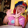 child, toddler, cap, backwards_cap, pink_shirt, indoor, playful, tongue_out, hand_gesture, person, young_child, furniture, ceiling_fan, light, home, portrait, cute, funny_face, expression, casual
