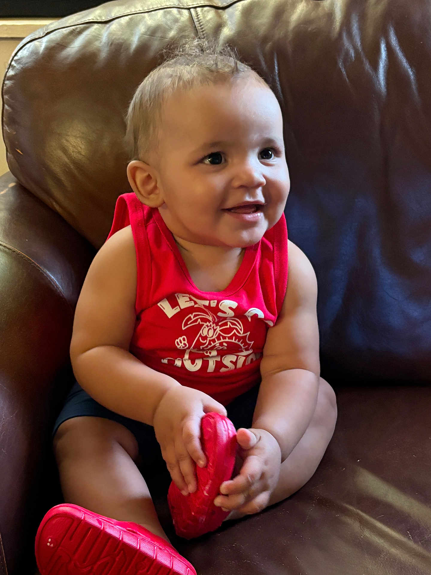 Hazen is registered to the contest to win money with this photo: baby, child, smiling, red_shoes, red_tank_top, sitting, leather_couch, indoors, cute, happy, person, portrait, infant, hand, footwear, furniture, casual_clothing, young_child, bright_colors, comfortable