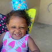 Journi Hall is registered to the contest to win money with this photo: blurred_background, casual, chair, child, colorful, curly_hair, cute, cute_expression, earrings, happy, indoor, person, playful, polka_dots, portrait, sleeveless_shirt, smiling, strawberry_pattern, toddler, young_child
