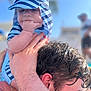 Archie is registered to the contest to win money with this photo: baby, child, striped_hat, striped_clothing, hand, adult, person, face, sun_hat, outdoor, blue_sky, water, wet_hair, skin, close_up, holding, summer, daylight, portrait, cute
